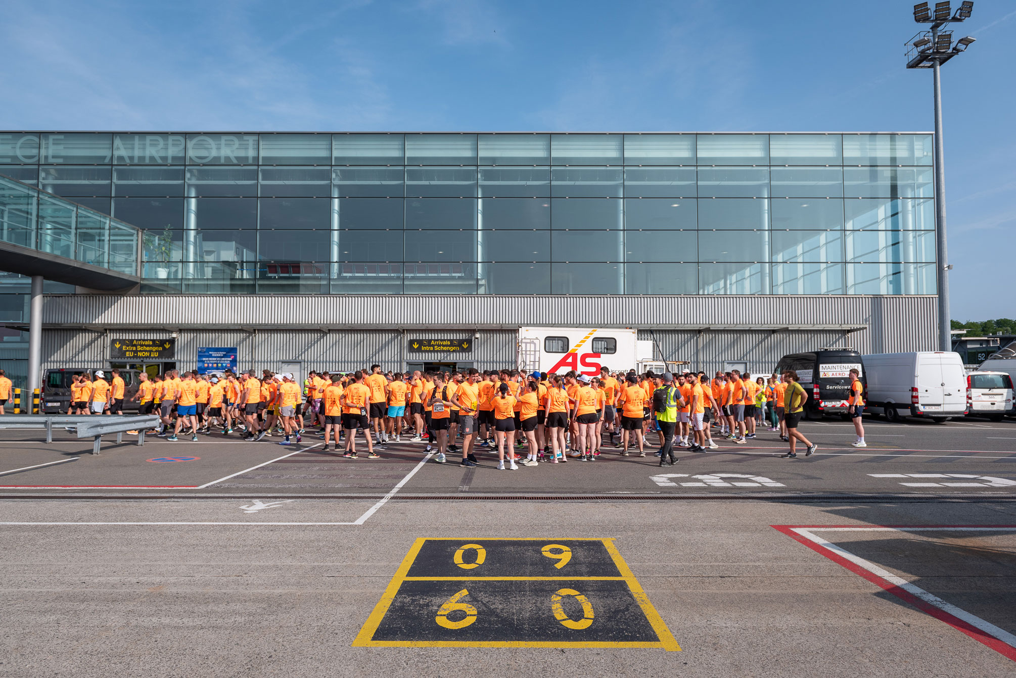 Une large foule de participants portant des t-shirts orange vifs est rassemblée sur le tarmac devant la façade vitrée de l'aéroport de Liège. On aperçoit des panneaux "Arrivals" et des marquages au sol jaunes typiques d'une zone aéroportuaire sous un ciel bleu clair.