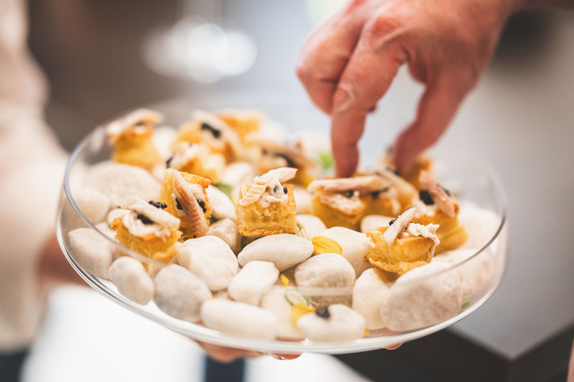 Gros plan sur un plateau de service transparent contenant des amuse-bouches gastronomiques disposés sur des galets blancs. Les bouchées se composent de petits carrés de gaufre surmontés d'une crème, d'un morceau de poisson fumé et d'une pointe de caviar. Une main s'apprête à en saisir un.