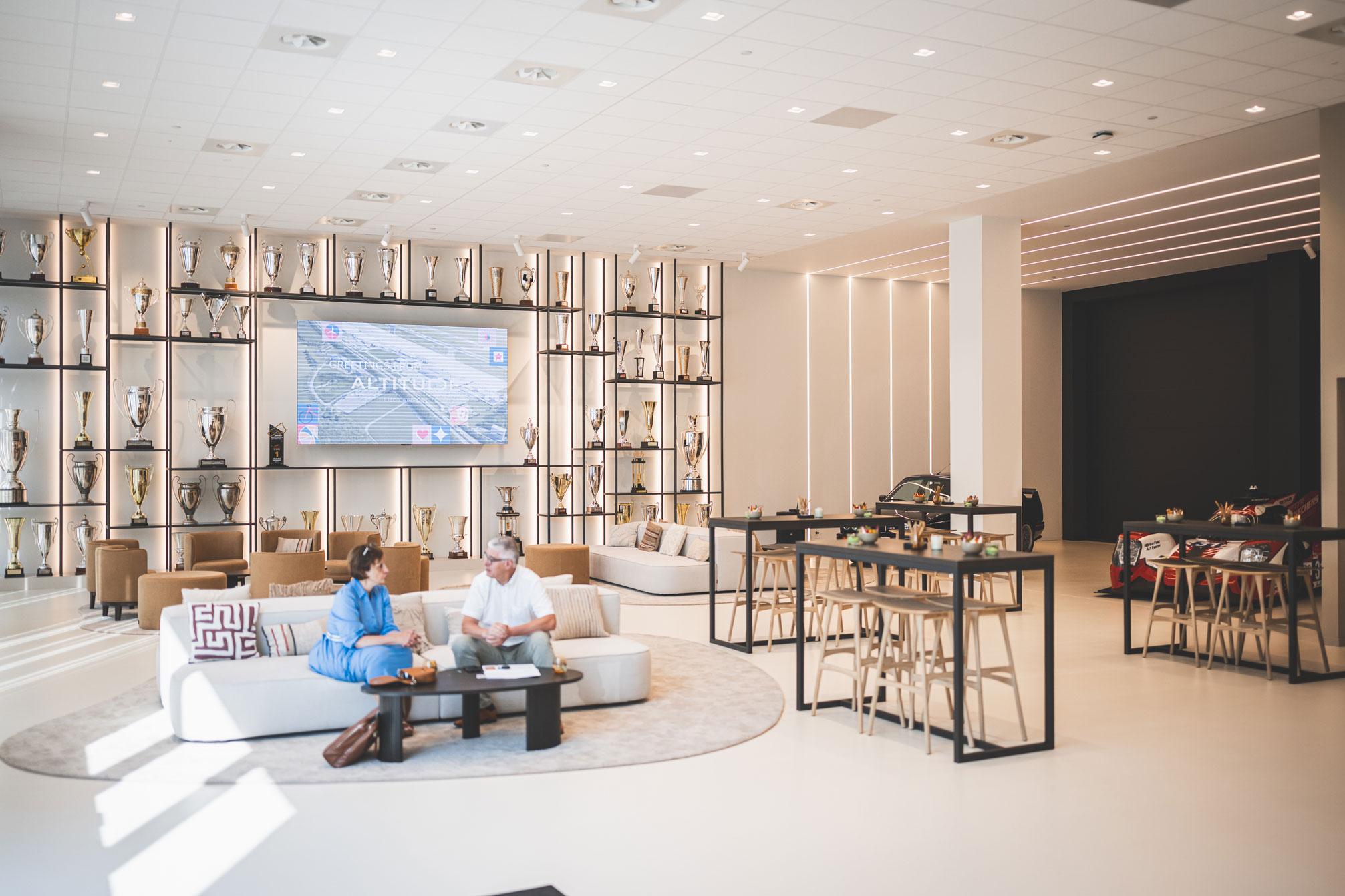 Intérieur d'un salon VIP moderne et lumineux à l'aéroport de Liège. Un mur entier est décoré de nombreuses coupes et trophées rétroéclairés entourant un écran plat. Au premier plan, un homme et une femme discutent sur un canapé blanc. Des tables hautes et une voiture de course sont visibles en arrière-plan.
