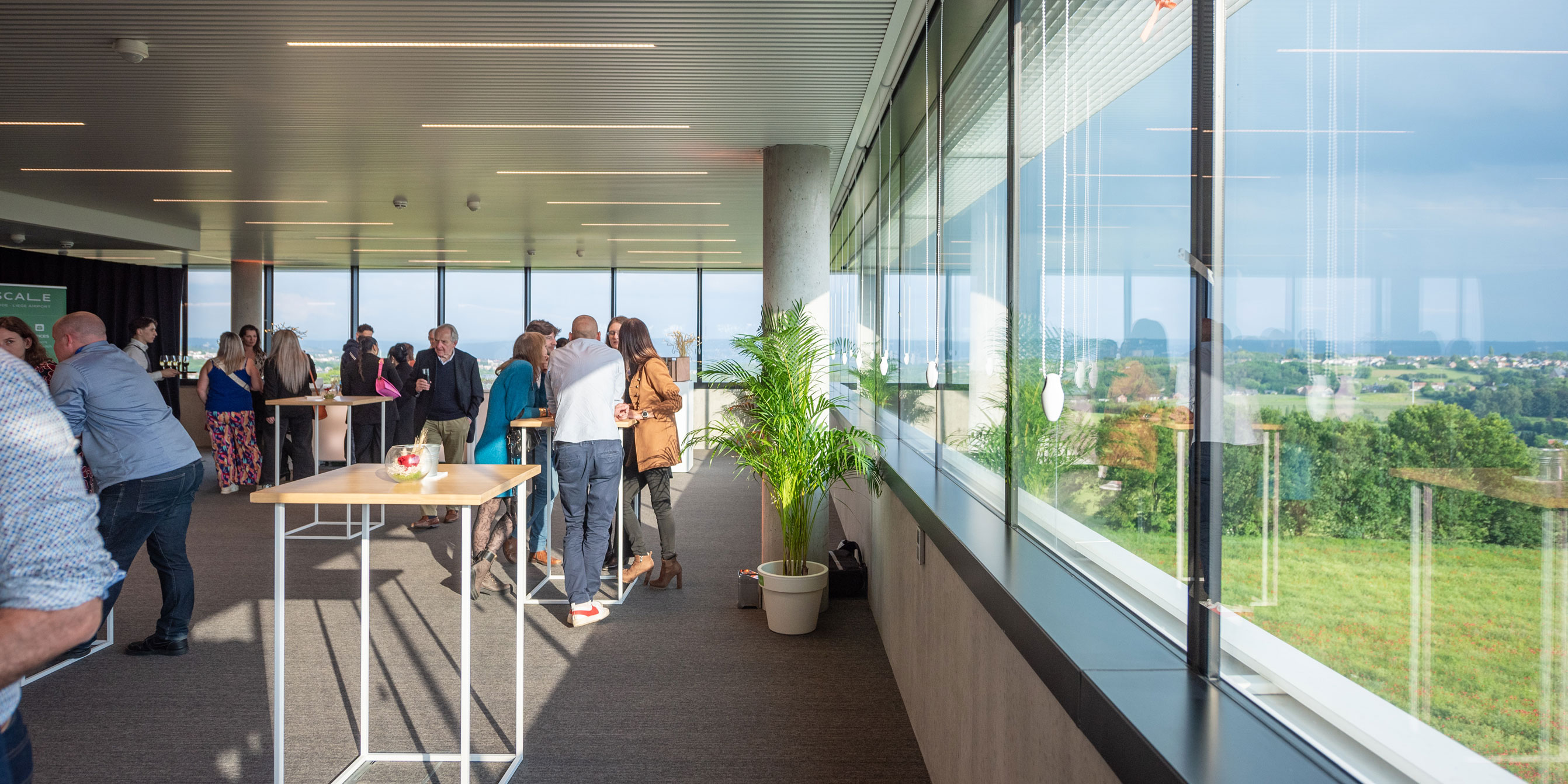 Une salle de réception spacieuse avec de grandes baies vitrées offrant une vue panoramique sur la campagne verdoyante. Des invités discutent debout autour de tables hautes. L'espace est baigné de lumière naturelle, créant une atmosphère de cocktail professionnel.