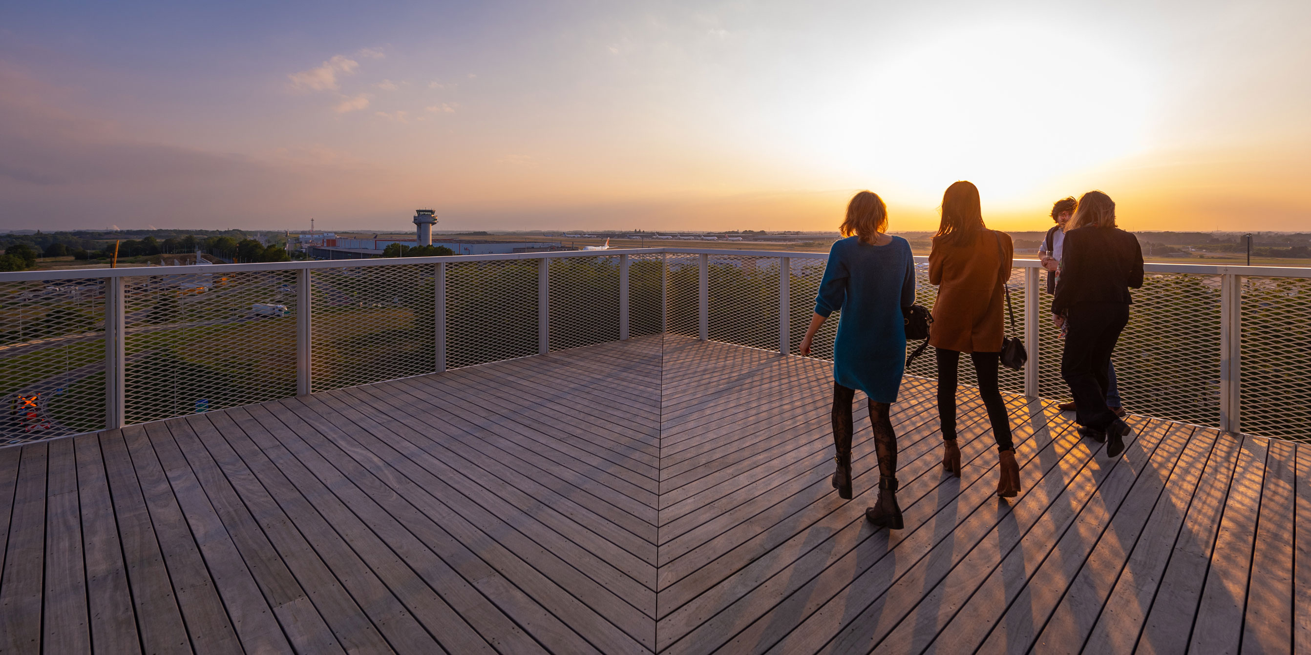 Des silhouettes de personnes marchant sur une grande terrasse en bois à l'heure dorée. La terrasse surplombe les pistes de l'aéroport de Liège, avec la tour de contrôle visible au loin. Le soleil couchant crée un contre-jour chaleureux sur l'horizon.